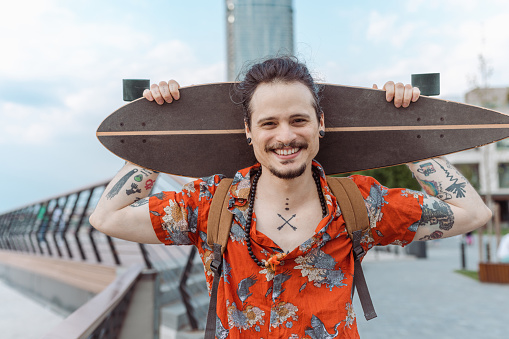 Portrait of a cheerful young man with a tattooed body carrying a skateboard on his shoulders and looking at the camera