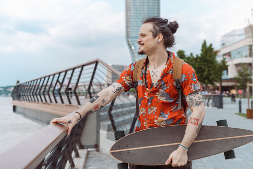 A handsome man with a tattooed body is holding a skateboard and looking at the river while standing in the downtown district. He is wearing casual clothes.