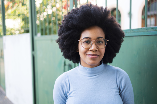 Portrait of a smiling Afro-Caribbean woman. She si wearing light blue blouse on a green background She is looking at camera