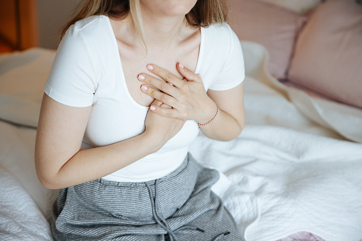 Heart attack, women with chest pain suffering at home, health problems concept. Young women with eyes closed holding his chest in discomfort, suffering from chest pain while sitting on bed at home. Elderly and health issues concept