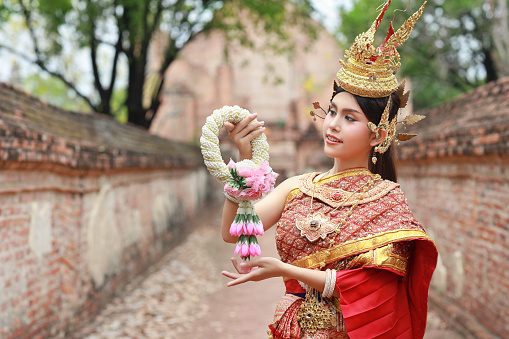 Young fashion and beautiful asian woman wearing Thai red traditional costume holding jasmine flower garland with happy and peace standing outdoor in ancient temple Ayutthaya, Thailand. Travel concept