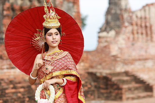 Young fashion and beautiful asian woman wearing Thai red traditional costume holding jasmine flower garland with happy and peace standing outdoor in ancient temple Ayutthaya, Thailand. Travel concept
