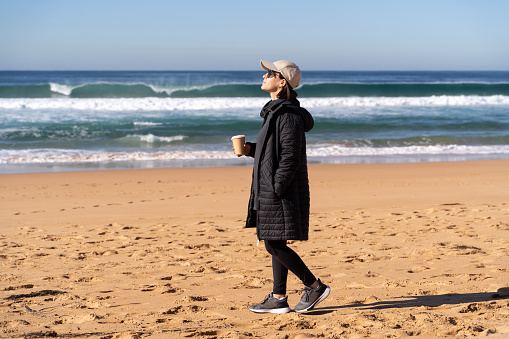 A woman walking with a cup of coffee in the winter beach, Australia, NSW