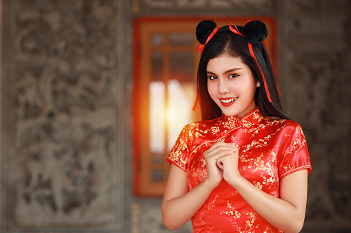 Beautiful young Asian woman in red Chinese dress traditional cheongsam qipao with gesture of congratulation and happy smiling face, she standing in front of the ancient wall (Chinese new year concept)