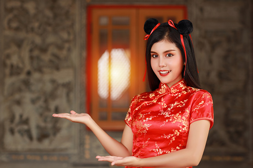 Beautiful Asian woman in red Chinese dress traditional cheongsam qipao with gesture of introduce or show something with happy smiling face, she standing in front of the ancient wall