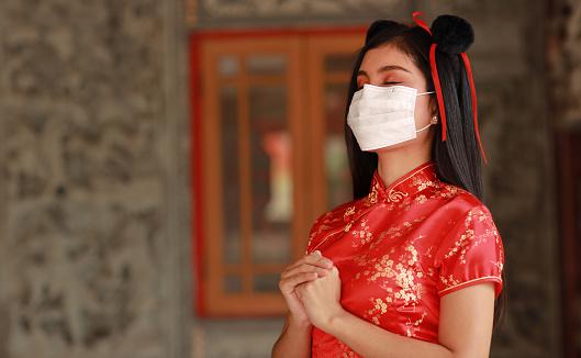 Beautiful young Asian woman in red Chinese dress traditional cheongsam qipao wear protect mask for healthcare with gesture of congratulation (New normal and Chinese new year concept)