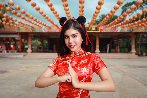 Beautiful young Asian woman in red Chinese dress traditional cheongsam qipao with gesture of congratulation and happy smiling face, she standing in front of ancient temple (Chinese new year concept)