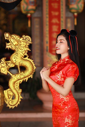 Beautiful young Asian woman in red Chinese dress traditional cheongsam qipao with gesture of congratulation and happy smiling face, she standing in front of ancient temple (Chinese new year concept)