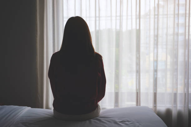 rear view image of a woman sitting alone on a bed in bedroom - depression women sadness window imagens e fotografias de stock