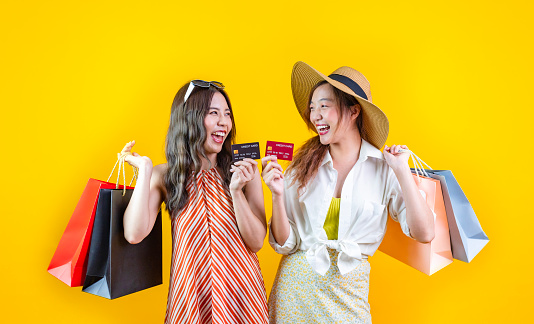 Portrait of two young asian women in casual beach clothing with credit card and shopping bag on sale promotion isolated on yellow background for travel and fashion studio cut out