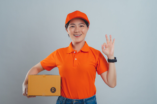 Asian woman in uniform of delivery person wearing orange polo t-shirt and holding box showing OK sign standing isolated over white background.