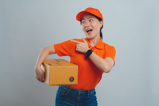 Asian woman in uniform of delivery person wearing orange polo t-shirt and holding box happy excited pointing to copy space standing isolated over white background.