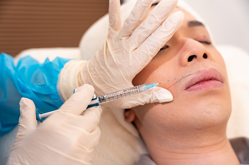 An young asian man is having a injection (or Aesthetic injection, filler injection, Hyaluronic acid injection , collagen buildup injection) in a beauty centre. He is lying on a bed and lying down patiently in a room.   A white towel is covering his head.  It is a close-up shot showing that the female doctor is performing the injection. she is wearing light blue healthcare uniform . She is holding the injector (syringe),  pointing at the man's  face with her hands in white gloves.