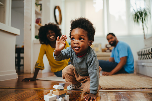 A playful happy african american child is playing a game with a Montessori train toy while his parents are laughing at him from the background. A happy child is having fun while learning from home.