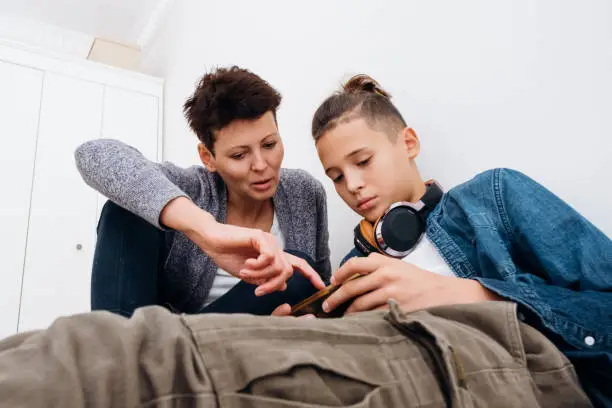 Photo of Teenager boy addicted to technology watching social video at home with his mother. Child and mother communicate and play with the technological device mobile phone