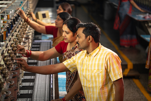 High angle view of male and female managers inspecting manufacturing equipment while workers producing spools in background at textile factory
