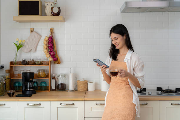 Young woman using smartphone leaning at kitchen table with coffee mug and modern home organizer Smiling woman reading phone message. stock photo