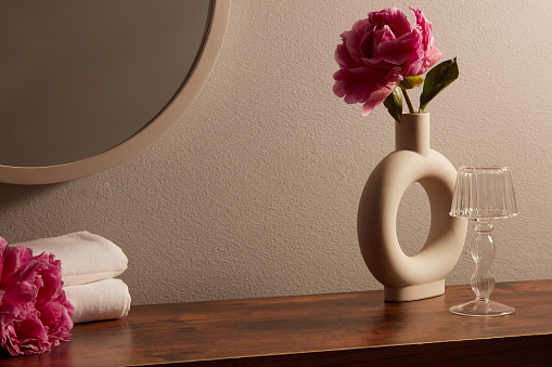 The interior decorations are placed on the wooden table - vase with pinks peonies, white towel and round mirror on brown background. Blank space for display your cosmetic product.