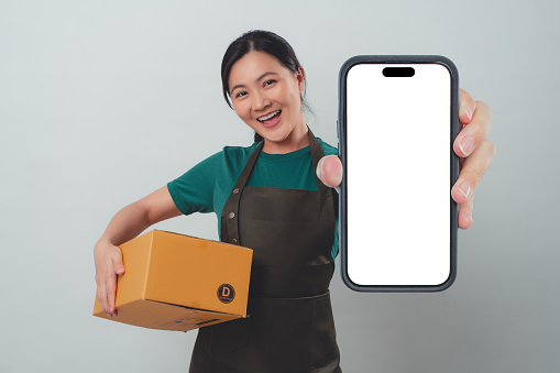 Asian woman wearing apron happy smiling holding and showing white screen on smartphone standing isolated over background.