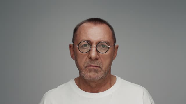 Portrait of Adult Man in Glasses Looking at Camera in Color Studio Shot. Old Male Isolated Alone on Grey Background Closeup. Aged Person Turning Head and Staring Focused with Normal Face Expression