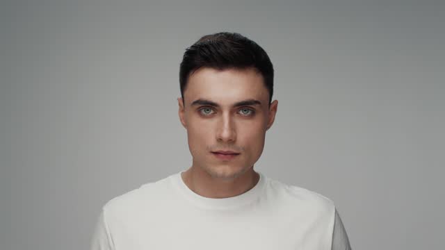 Portrait of Young Man Looking at Camera in Color Studio Shot. Adult Boy Isolated Alone on Grey Background Close-up. Beautiful 20s Person in Trendy t-shirt Raising Head and Opening Bright Pretty Eyes