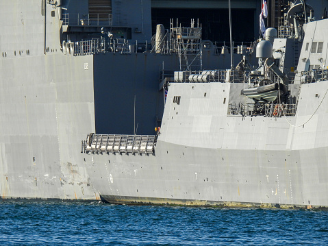 The sterns of HMAS Brisbane (right) and HMAS Canberra (left) of the Royal Australian Navy.  They are docked at Garden Island, Sydney Harbour.  The serial number of each ship is painted on the far stern. HMAS Brisbane is one of three Hobart Class destroyers in the fleet.  HMAS Canberra is one of two Amphibious Assault Ships in the fleet.  This image was taken on a sunny afternoon on 20 May 2023.