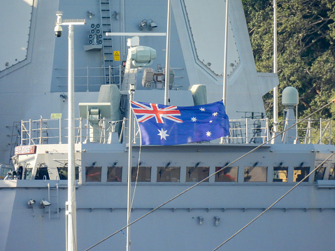 The bridge and superstructure of HMAS Sydney, a Hobart Class destroyer of the Royal Australian Navy.  She is docked at Garden Island, Sydney Harbour. The flag is attached to a flagpole on the bow of HMAS Brisbane, docked next to her.  A post with security cameras on the dock has spikes on the top to deter birds.  This image was taken from Woolloomooloo Bay on a sunny afternoon on 20 May 2023.