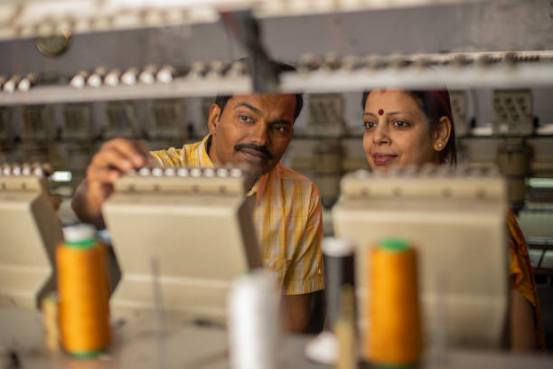 Mid adult focused Indian male and female workers holding threads in spool manufacturing machinery at textile factory
