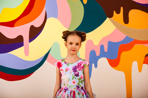 Happy adorable little girl in colorful flowers dress at multi colored wall, smiling looking at camera. Stylish child pretty model posing, studio shot. Fashionable kid style concept. Copy ad text space