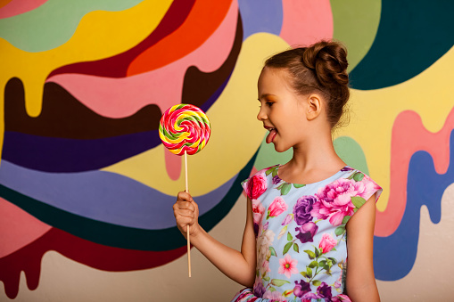 Playful little girl 6-7 year old stuck out her tongue, looking on round lollipop at multi colored wall. Lovely kid in colorful flowers dress with candy on stick. Summer sweet concept. Copy text space