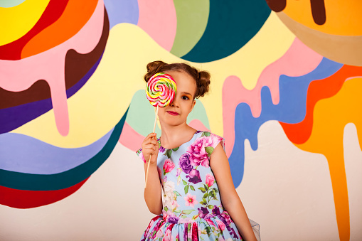 Little girl 6-7 year old covering eye with round lollipop at multi colored wall, smiles looking at camera. Cute child in colorful flowers dress with candy on stick. Summer sweet concept. Copy ad space
