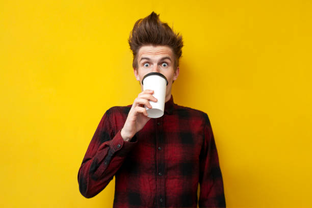 shocked guy drinks invigorating coffee and is surprised at a yellow isolated background, a man drinks an energy drink