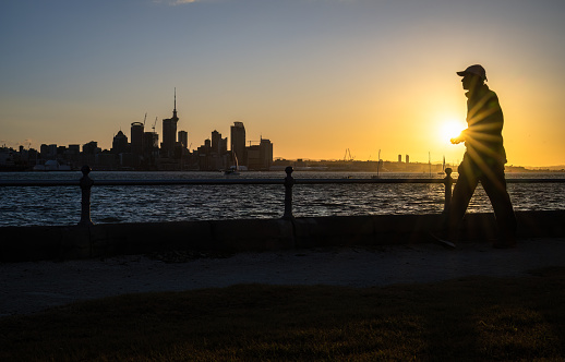 Man walking at Devonport waterfront at sunset. Skytower and Auckland city high-rises in the background.