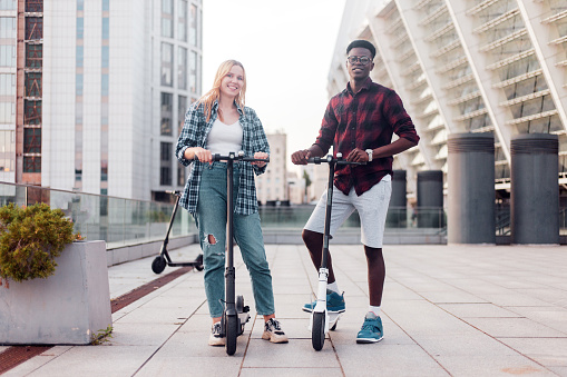 interracial young couple with scooters on the city background, european girl and african american guy ride scooters and smile, eco transport rental