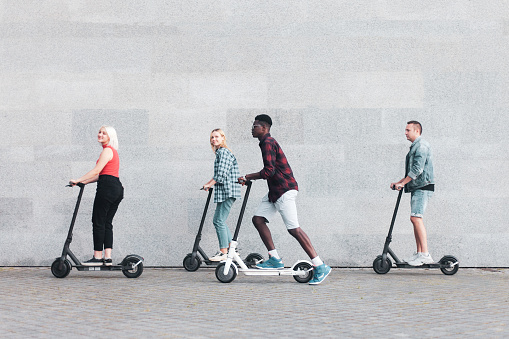 interracial company of friends rides electric scooters against the background of a wall, young people use eco transport in the city