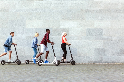 interracial company of friends rides electric scooters against the background of a wall, young people use eco transport in the city
