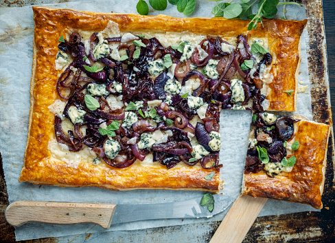 Overhead view of a large red onion tart. Made with caramelised red onions, blue cheese or gorgonzola cheese and oregano. Delicious as an appetiser or a light lunch or brunch. Colour, horizontal format with some copy space. 
Made with shop bought puff pastry. Roll out your pastry and make a gentle cut about 2cm in for the edge and cut all the way around  the pastry being careful not to cut through, brush this edge with a beaten egg. Inside this prick the pastry base with a fork to stop it for rising when it is baked, pre-bake this for 10 mins. Make your caramelised onions by peeling and cutting the red onions into rings. Melt some butter and fry the onions until they are soft, add sugar, balsamic vinegar and simmer for between 10-15 minutes.
Then spread this mixture over the pre-baked pastry base with some grated cheese, mozzarella, and some gorgonzola cheese, or similar, add some fresh oregano and bake for 10-12 minutes. Cool on a wire rack, enjoy!