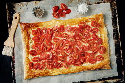 Puff pastry tart with tomatoes, cheese and fresh oregano. Delicious as an appetiser or a light lunch or brunch. 
Colour, horizontal format with some copy space. 
Made with shop bought puff pastry. Roll out your pastry and make a gentle cut about 2cm in for the edge and cut all the way around  the pastry being careful not to cut through, brush this edge with a beaten egg. Inside this prick the pastry base with a fork to stop it for rising when it is baked, pre-bake this for 10 mins. 
Start making the filing by halving the cherry tomatoes and remove some of the moisture in them by placing them face down on some kitchen paper for 20 minutes or so . Mix 3 egg yolks with 150g of sour cream, 150g of grated cheese and a spoonful of dijon mustard.
Then spread this mixture over the pre-baked pastry base and place the tomatoes cut side up on top of the mixture, add some fresh oregano and bake for 10-12 minutes. Cool on a wire rack, enjoy! Serve with some fresh watercress salad leaves.