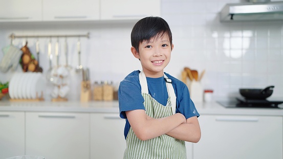 Confident asian little boy wearing apron smiling to camera whlie standing with arms crossed in kitchen room at home. Dream career concept