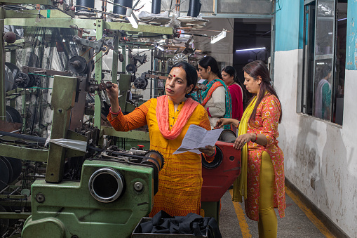 Mature woman with paper inspecting manufacturing equipment and female workers working at production line in textile factory and representing women empowerment