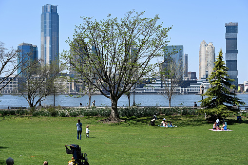 New York, USA, April 13, 2023 - Tourists and locals relax in Hudson River Park in Lower Manhattan on a warm spring day. In the background the Hudson River and the skyscrapers of Hoboken, New Jersey.