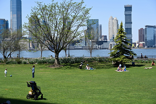 New York, USA, April 13, 2023 - Tourists and locals relax in Hudson River Park in Lower Manhattan on a warm spring day. In the background the Hudson River and the skyscrapers of Hoboken, New Jersey.