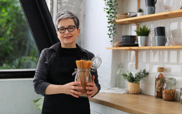 femme âgée souriante portant des lunettes et un tablier noir tenant des spaghettis crus dans la cuisine dans ses mains. - eating senior adult color image spaghetti photos et images de collection