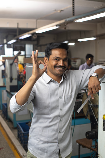Portrait of smiling male manager showing ok sign gesture while standing by manufacturing equipment in textile factory