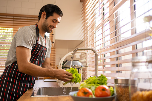 Cheerful young Hispanic man in grey shirt and apron washing vegetables tomatoes on the kitchen.