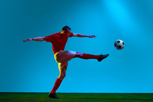 Hitting ball with leg. Portrait of soccer, football player in sports uniform kicking the ball in motion over field background in neon light. Concept of team game, sport, championship, competition, ad