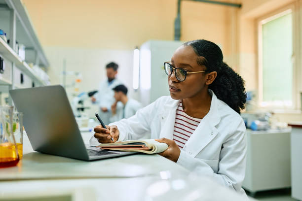 istock Black female biochemist writing research data in notebook while working on laptop in laboratory. 1493124934