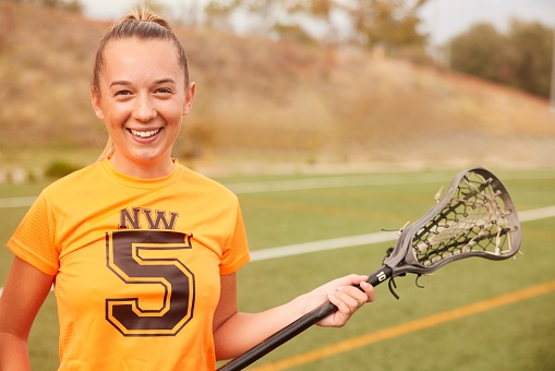 A lacrosse female player looks directly at the camera with a joyful smile on her face. The waist-up view captures her positive attitude and enthusiasm for the game.