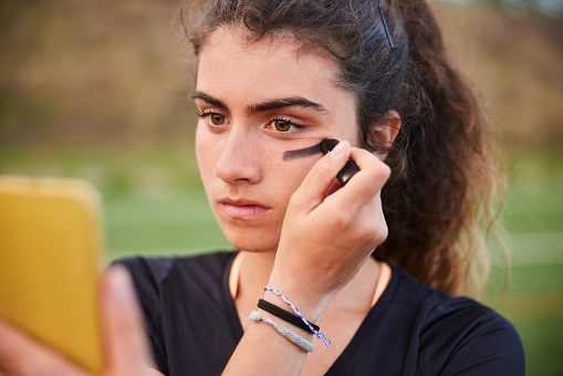 A lacrosse female player uses her smartphone as a mirror to apply eye black on her face.
