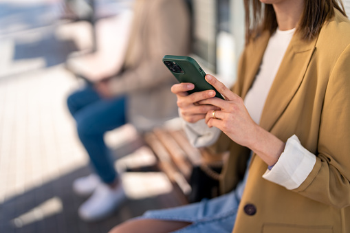 Unrecognizable modern young woman holding mobile phone while sitting at bus station and waiting for bus, using smart phone to check bus schedules, connections and maps, buying bus ticket on online app.